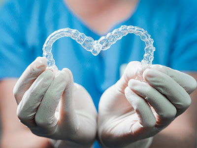 The image shows a hand holding a clear plastic heart with a pair of transparent braces inside it, placed on a blue surgical glove against a white background.