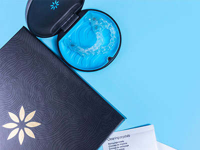 The image shows a blue toothbrush with a clear case containing crystals, placed next to a black box with a white logo on a light blue background.