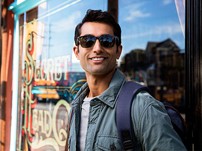 The image shows a man wearing sunglasses, standing outdoors with a backpack, smiling at the camera. He is positioned in front of a storefront window displaying a sign that reads Sweet Treats.